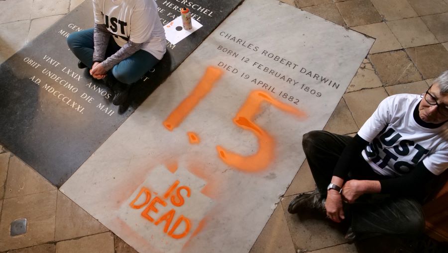 Caption: A handout picture taken and released by the Just Stop Oil climate campaign group on January 13, 2025, shows two activists, sitting on the floor alongside the grave of Charles Darwin, after they sprayed "1.5 is dead" over it in protest against climate change in action, inside Westminster Abbey in central London. Credit: AFP PHOTO / JUST STOP OIL / JAMIE LOWE 